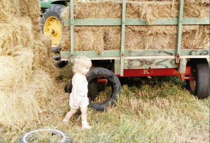 Hicks, Hoodlums, & Highrises author, Nicole Braun, as a toddler walks behind a full wagon of bailed hay