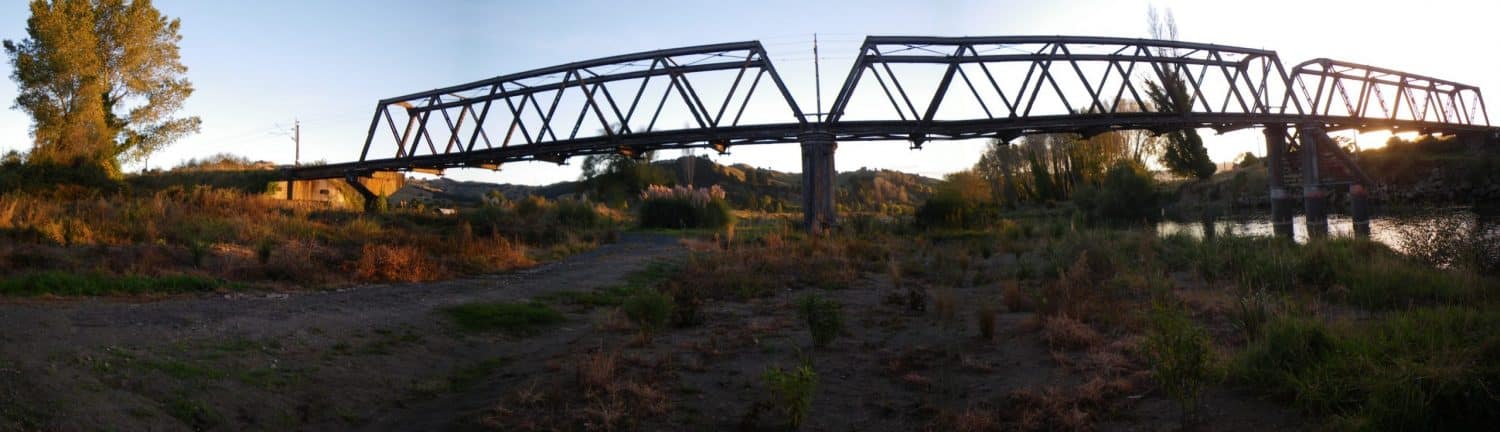 Sun-setting behind a iron railway bridge over a stream and gravel road in Taumarunui, New Zealand
