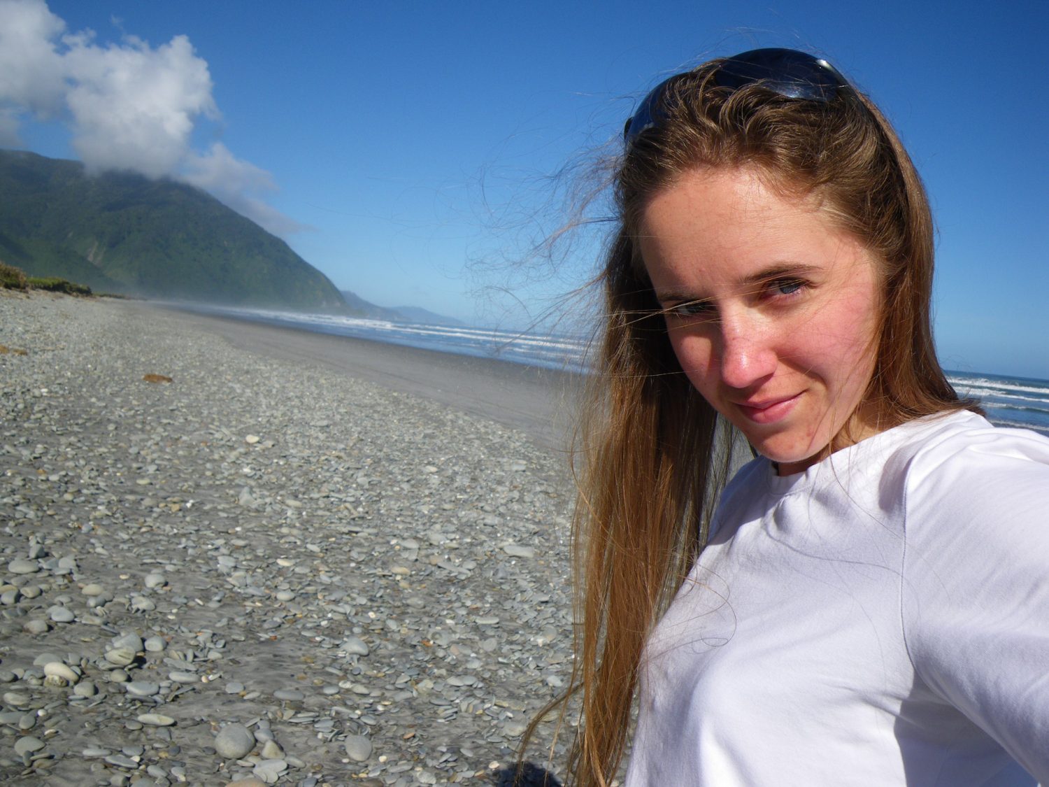 Hicks, Hoodlums, & Highrises author, Nicole Braun, snaps a selfie on a pebble lined beach in Barrytown, New Zealand with mountains cascading into the ocean in the background