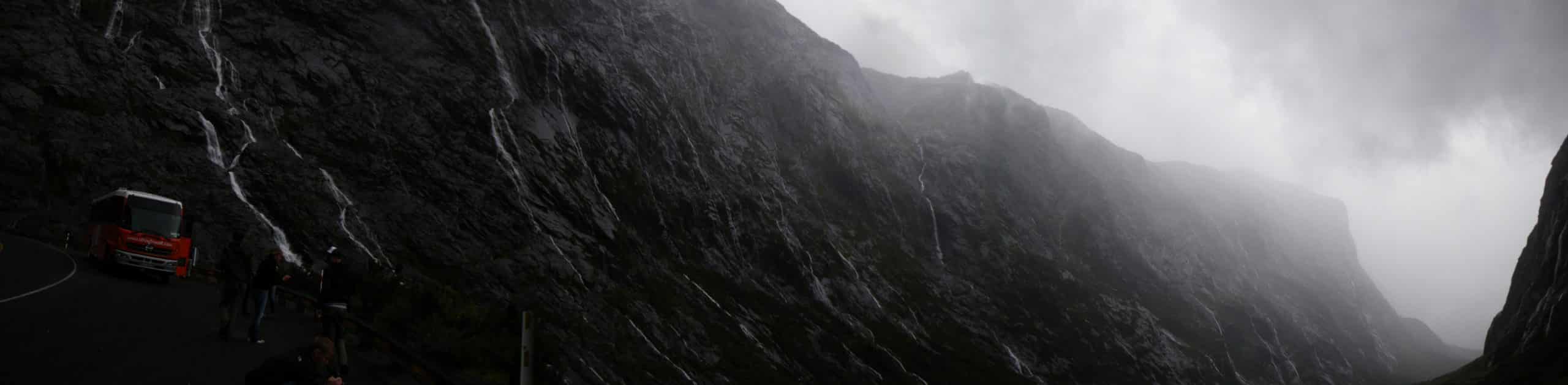 Panoramic image of black stone valley walls with spindly waterfalls cascading throughout as grey clouds ascend in the Valley of a Thousand Waterfalls, New Zealand