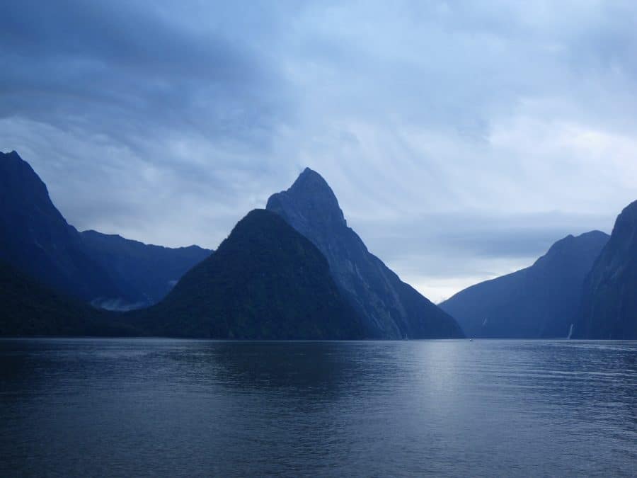 Calm water with steep, forested mountains under a cloudy sky coverd in a dark blue hue as a strom rolls into Milford Sound, New Zealand