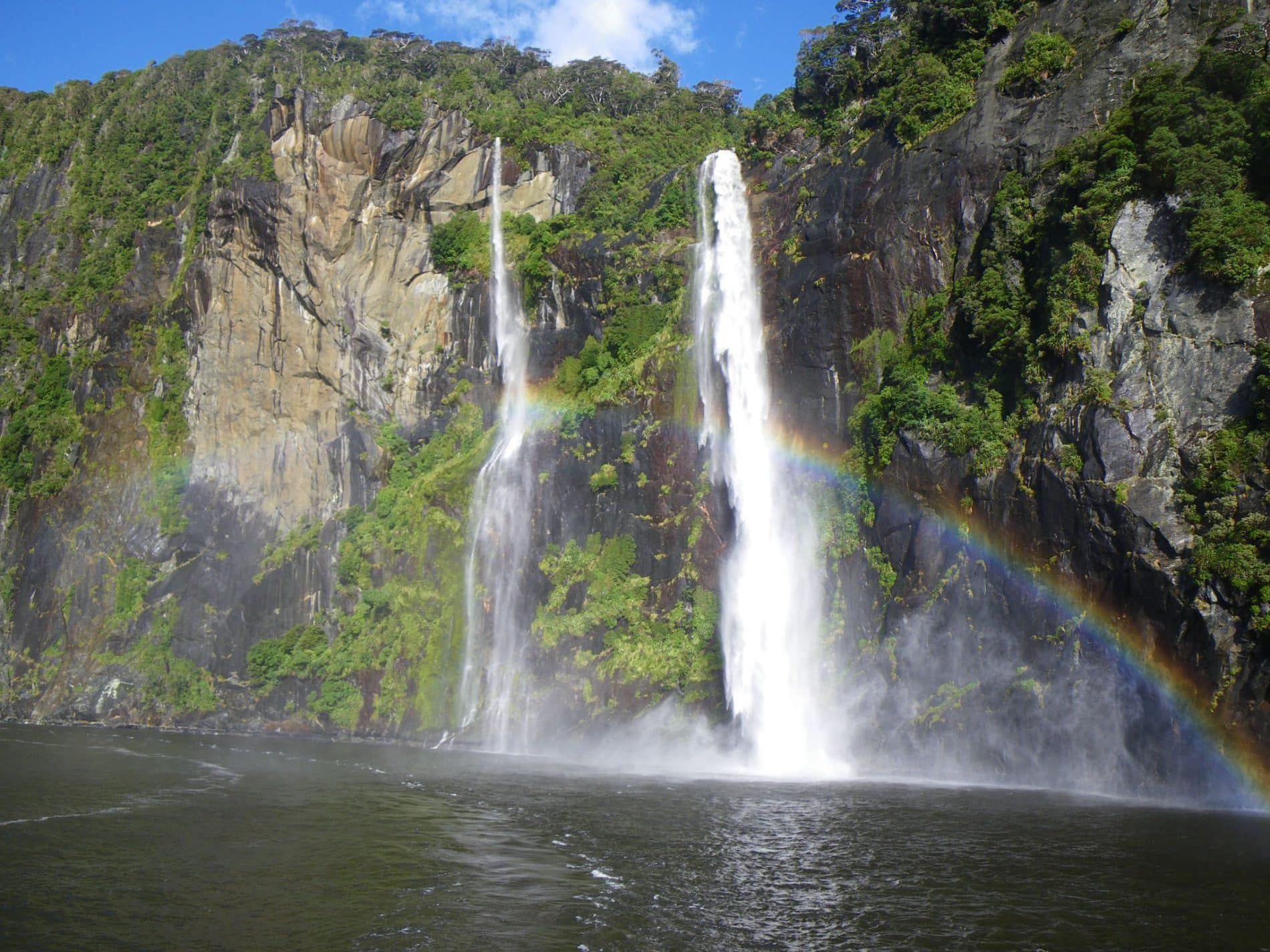 Two tall waterfalls cascade down a steep, green cliff into a river, with a clear rainbow forming in the mist at the base of the falls.