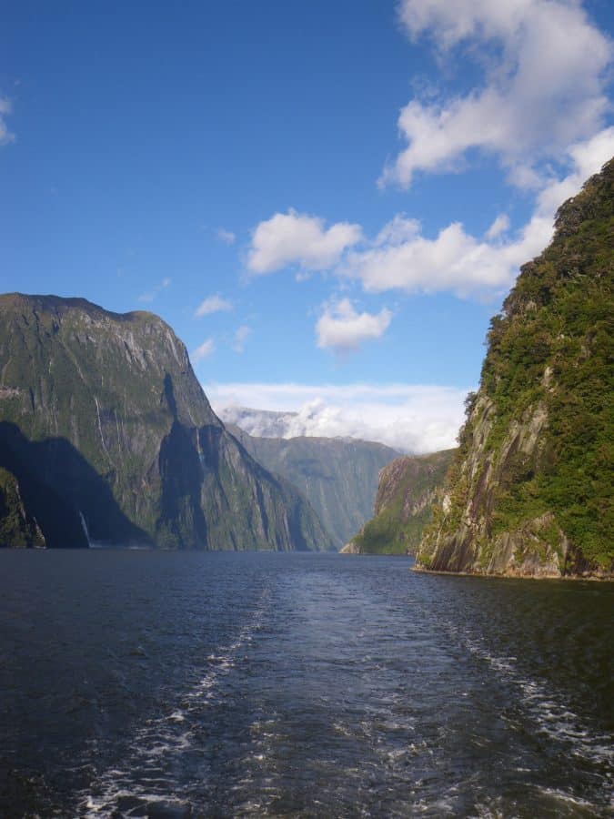 A view of a wide fjord with steep, green cliffs on both sides, a waterfall in the distance, and a boat wake in the water under a blue sky with scattered clouds, Milford Sound, New Zealand