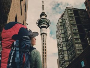 Hicks, Hoodlums, & Highrises author, Nicole Braun wearing a backpack, looks up at Auckland, New Zealand skyscrapers